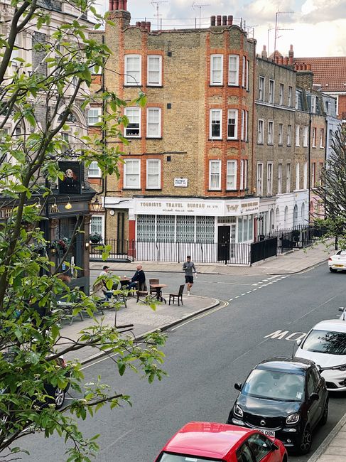 A street view showing a three-story brick building with large windows, situated on a corner at the intersection of King Street in Twickenham. The ground floor has a white shopfront with closed metal security shutters and a sign reading 'Torrents Travel Bureau.' In front of the building, there are black metal railings, and outside, two people are seated at a wooden table with benches, while another person stands nearby. Several cars, including a black hatchback and a red vehicle, are parked along the street. The scene includes some trees with green leaves partially visible in the foreground, and the daylight is natural with a slightly cloudy sky, illustrating a typical urban setting where home relocation or furniture transport logistics might be coordinated by services such as Twickenham Man and Van during a house move or moving project.
