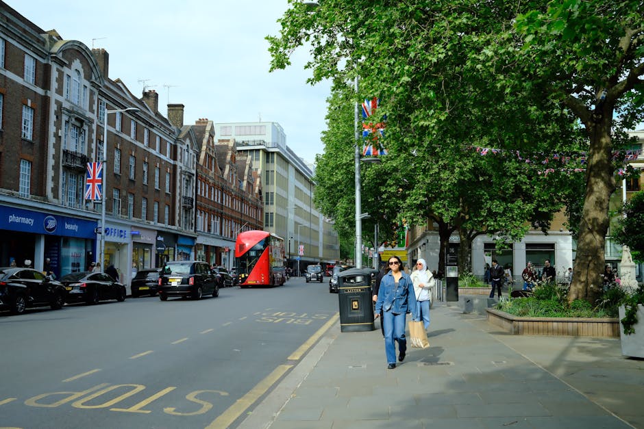 A busy street scene in Twickenham with a row of traditional brick and modern buildings lining the sidewalk, some with Union Jack flags displayed. Several parked cars, a red double-decker bus, and moving vehicles are visible along the road. The sidewalk features large green trees providing shade, with pedestrians walking, including two women in blue and white outfits carrying shopping bags. The area is well-maintained with planters, a trash bin, and street furniture. This setting is typical for a home relocation or moving logistics context, highlighting urban street access and movement. The image is associated with Twickenham Man and Van’s house removal services, emphasizing street access considerations for local furniture transport and packing and moving operations.