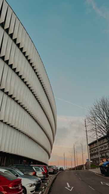 The image shows the exterior of a large, modern, curved building with metallic horizontal slats forming its facade, set against a partly cloudy sky during either sunrise or sunset. In the foreground, there is a parking area with multiple parked cars arranged along the curb, including sedans and hatchbacks in various colours. To the right of the parking lot, a narrow street extends into the distance, with a few trees without leaves indicating a winter or early spring setting. A utility pole with power lines runs alongside the street, and another building with multiple windows is visible further down the road. This urban scene is consistent with a location near Twickenham Stadium, where professional removals by Twickenham Man and Van could facilitate home relocations or furniture transport within the area.