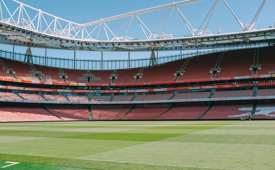 Photograph of the interior of a large football stadium with a well-maintained grass pitch in the foreground, and multiple tiers of red seating visible on three sides. The stadium has modern white structural supports and a partially open roof with visible trusses. The seating areas include sections with light pink, dark red, and black seats, and some advertising banners are displayed along the middle tier. The sky above is clear with a few clouds, and bright sunlight illuminates the scene. This image showcases the spacious environment of a professional sports venue, relevant to the context of house removals or transportation logistics in and around Twickenham, as seen on the Twickenham Man and Van website.