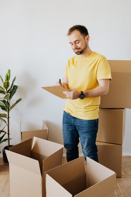 A man wearing a yellow t-shirt and blue jeans stands inside a residential property, holding a clipboard and pen, seemingly inspecting or preparing for a home relocation. In front of him are several op